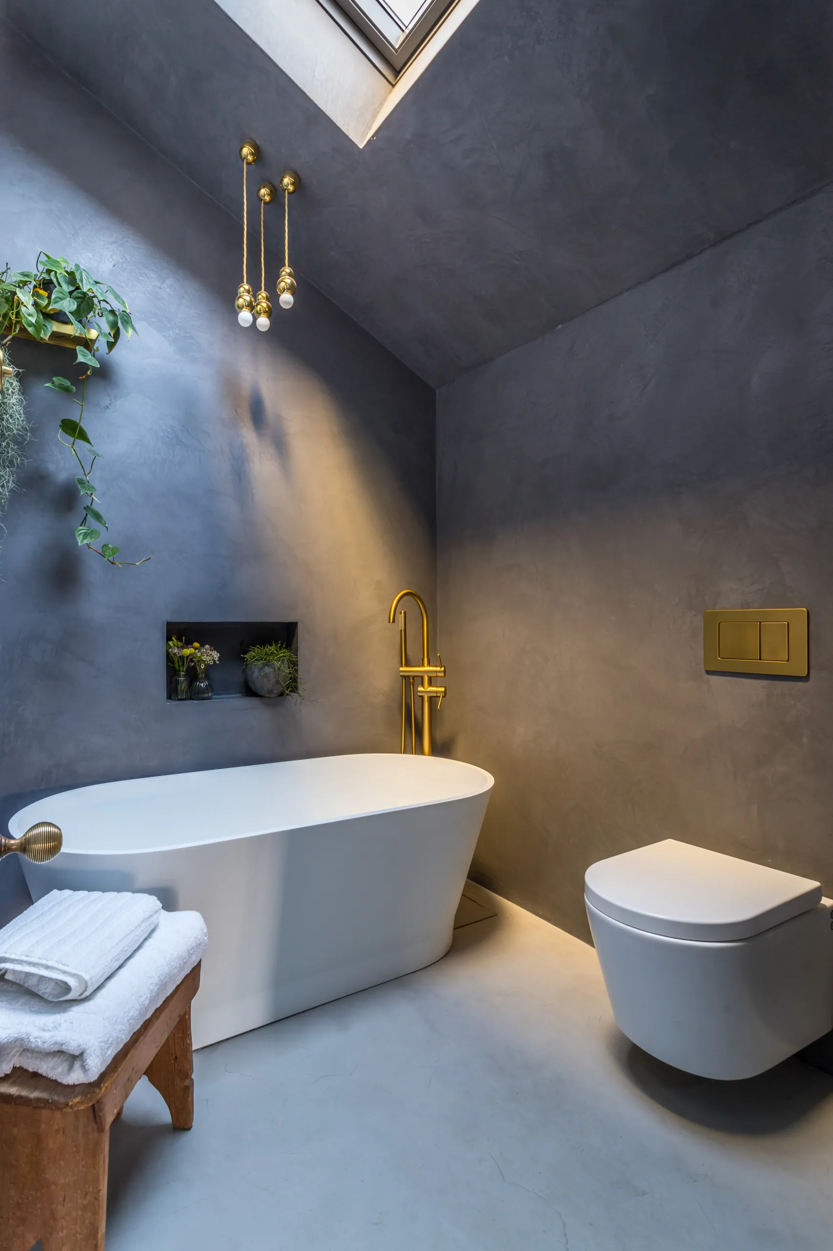 Bathroom in charcoal microcement: walls, sloped ceiling and floor run seamlessly around a freestanding tub, with brass fittings and a skylight.