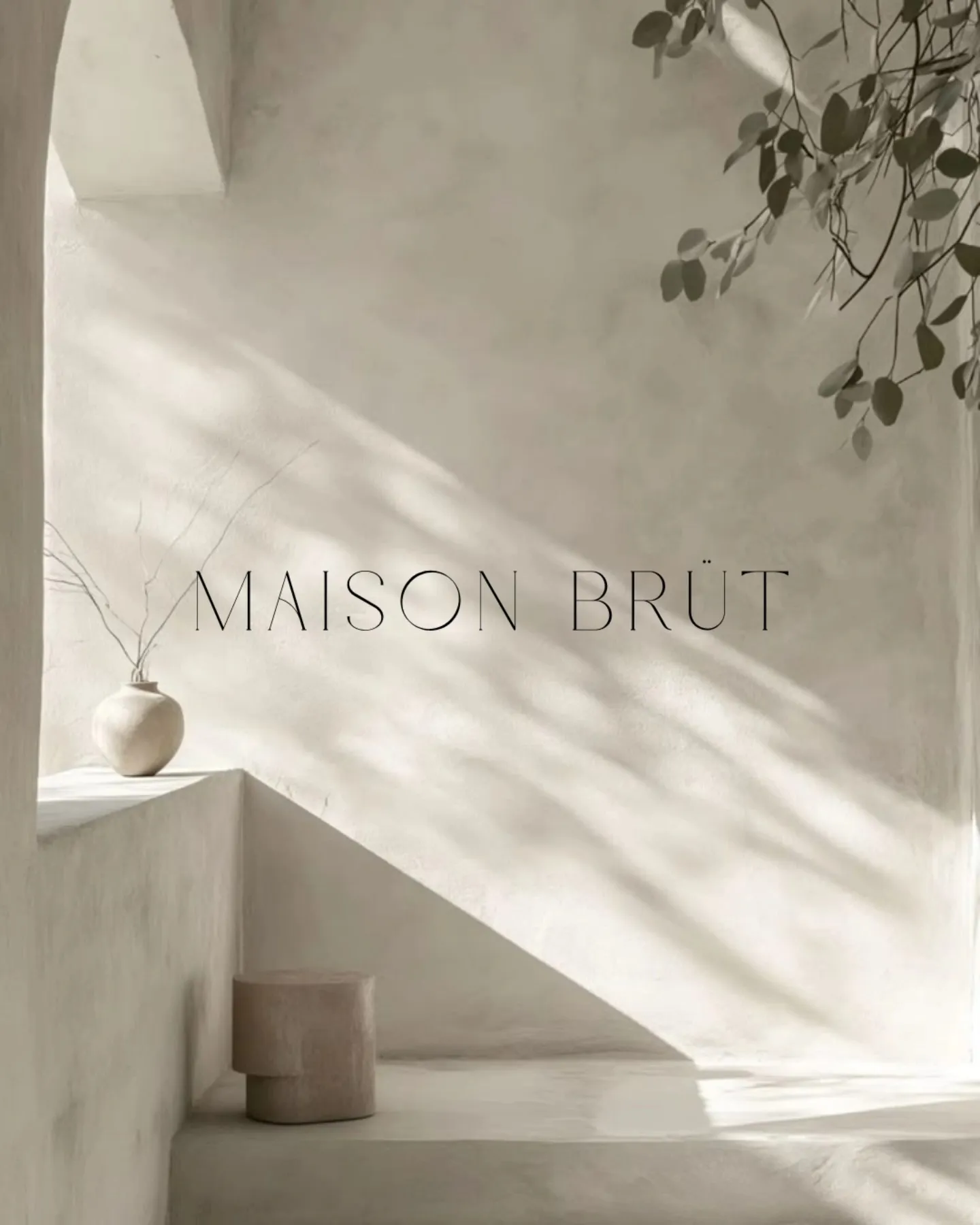 A plastered alcove in raking daylight, a pale vase on a built-in ledge and a cylindrical stool below, with the Maison Brüt wordmark set across the wall.