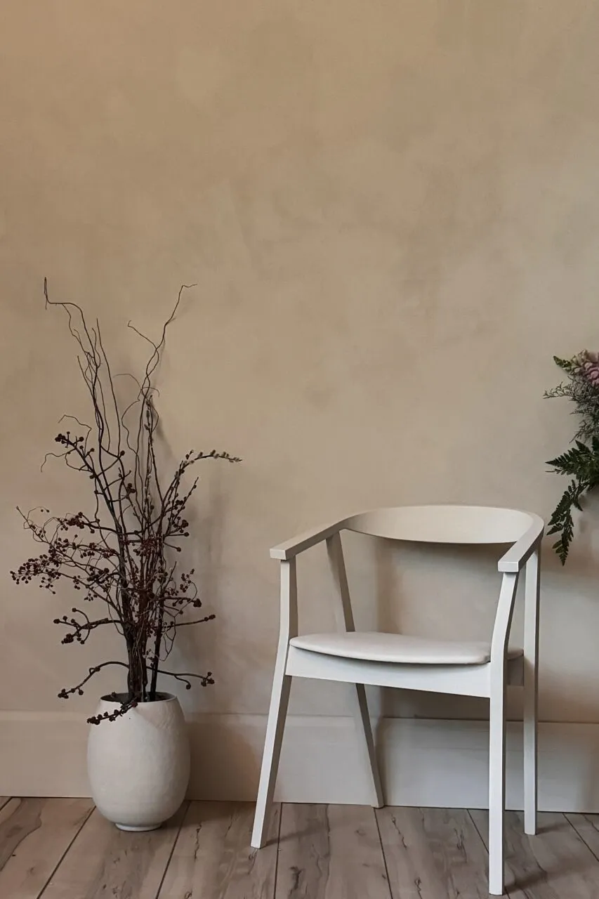 Wall in warm sand Venetian plaster, its soft mineral movement behind a pale wood chair and a stoneware vase of dried branches.